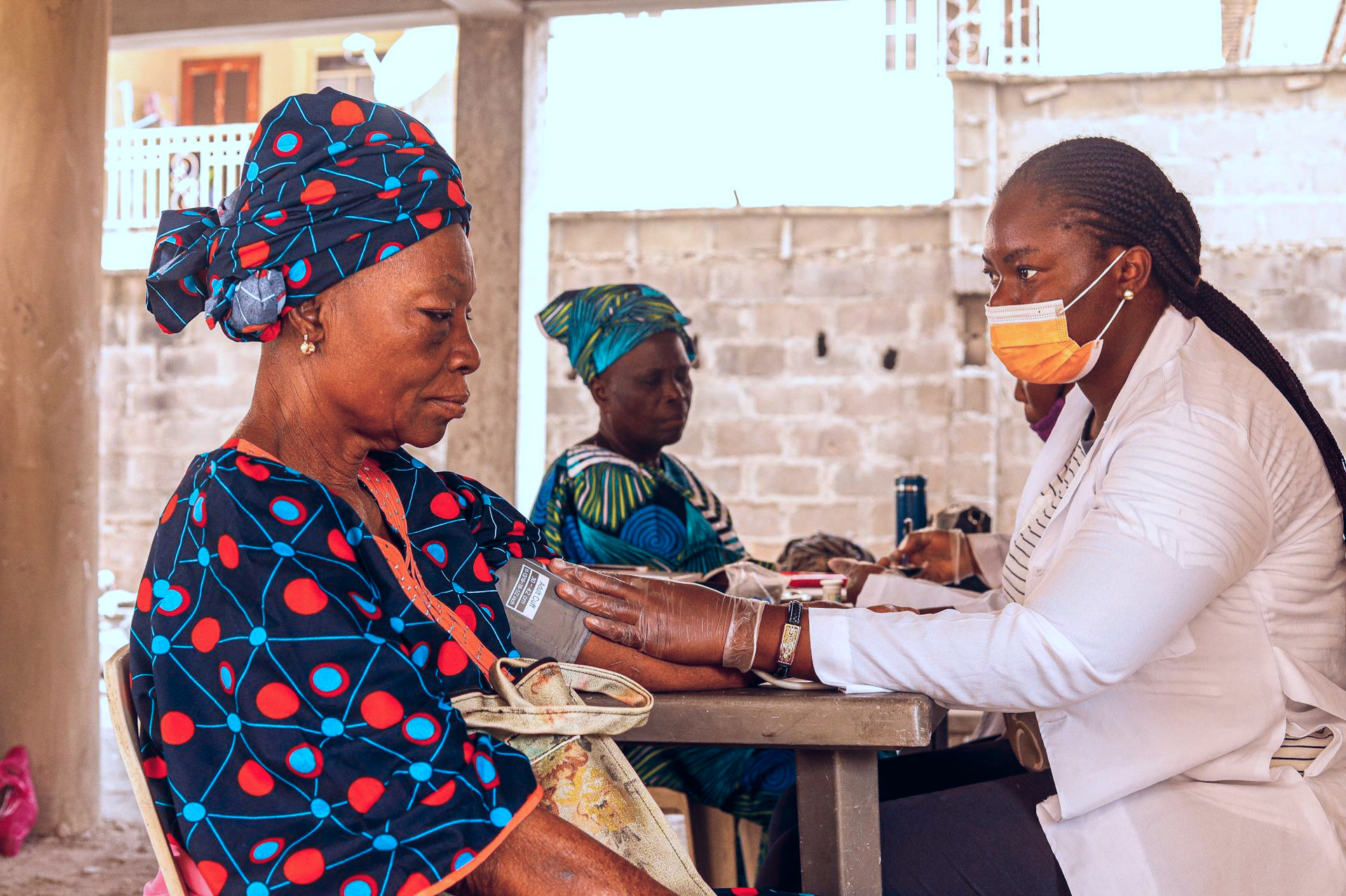 A healthcare worker wearing a white coat, gloves, and a face mask checks the blood pressure of an older woman dressed in a colorful outfit and headwrap.