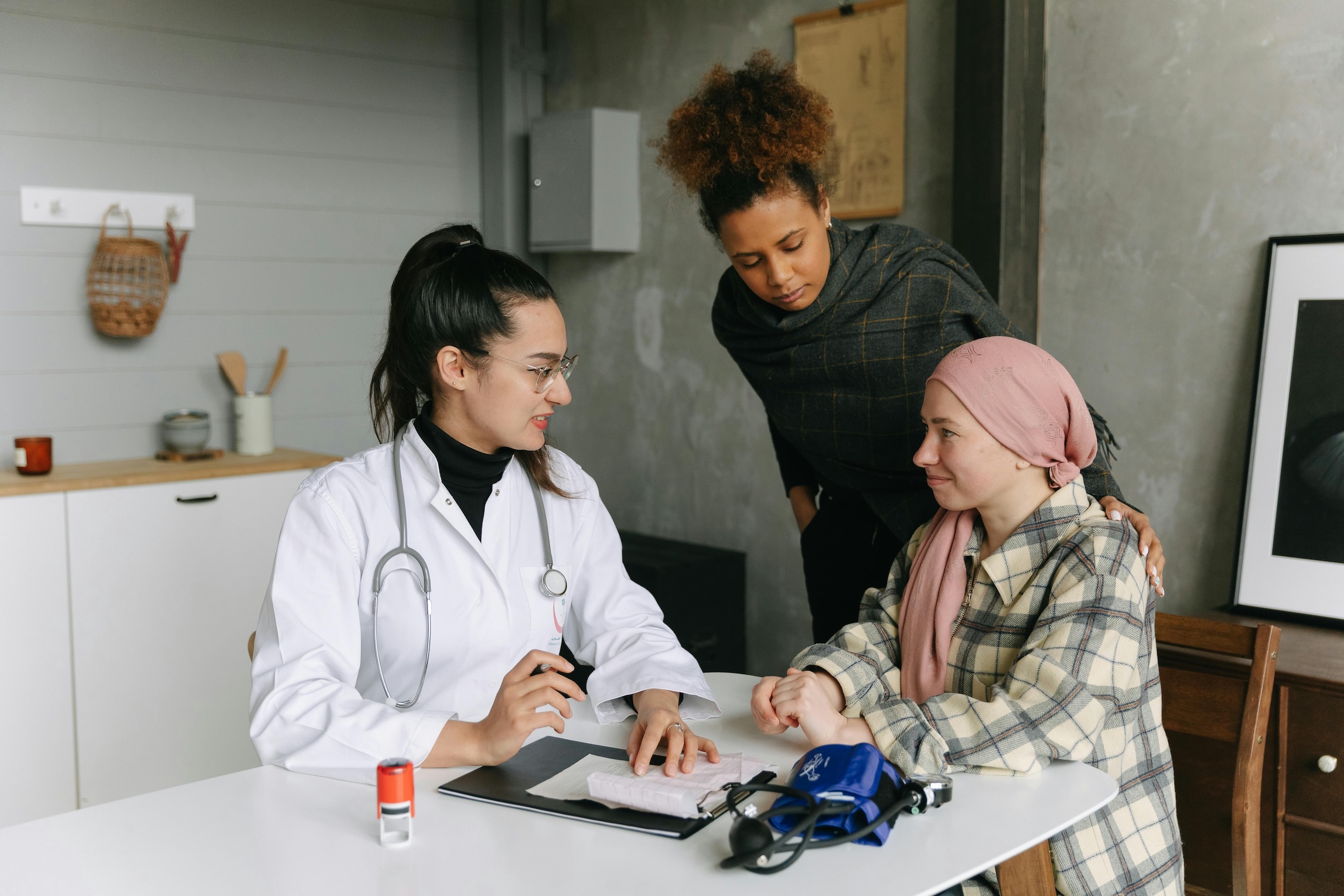 A female doctor in a white coat and stethoscope sits at a table talking with a patient wearing a headscarf and plaid jacket. Another woman stands beside the patient, offering support.