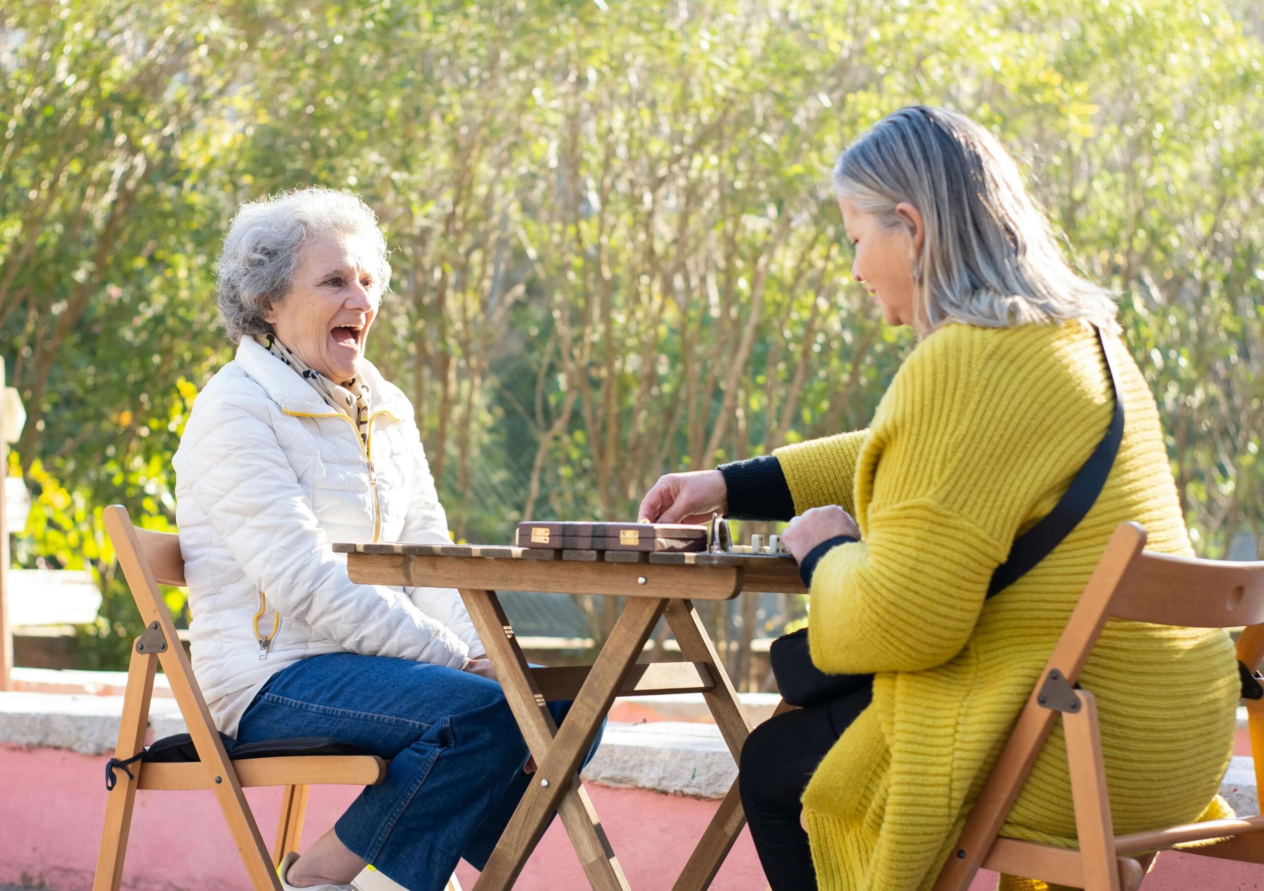 Two ladies sit happily across from each other at an outdoor table playing dominoes.