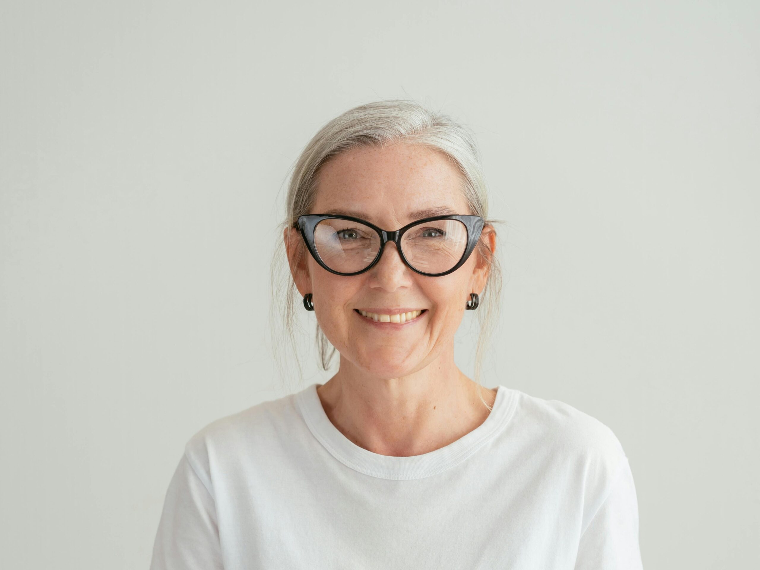 A smiling older woman with gray hair, black-framed glasses, and a white t-shirt stands against a plain light-colored background.