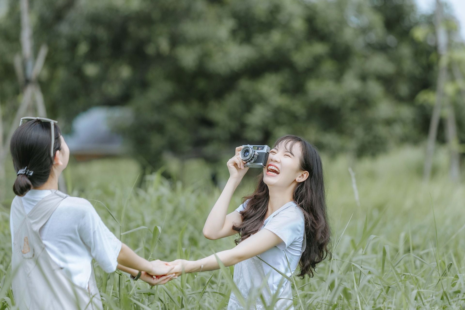 Two young women in a green field, laughing and holding hands, one has a vintage camera in her hand